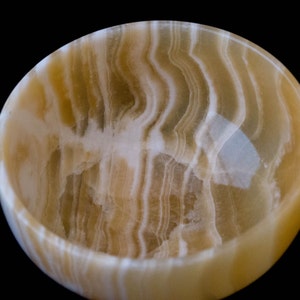 K&ouml;nnte beinhalten: Runde Schale aus Onyx mit einem nat&uuml;rlichen, wirbelnden Muster aus cremewei&szlig;en und goldbraunen Farbt&ouml;nen. Der Innenraum ist glatt und poliert. Die Schale steht vor einem schwarzen Hintergrund. Ideal f&uuml;r Dekorationszwecke.