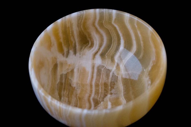 K&ouml;nnte beinhalten: Runde Schale aus Onyx mit einem nat&uuml;rlichen, wirbelnden Muster aus cremewei&szlig;en und goldbraunen Farbt&ouml;nen. Der Innenraum ist glatt und poliert. Die Schale steht vor einem schwarzen Hintergrund. Ideal f&uuml;r Dekorationszwecke.