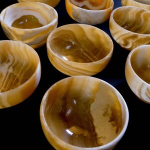 K&ouml;nnte beinhalten: Eine Sammlung kleiner, runder Schalen aus durchscheinendem, bernsteinfarbenem Stein. Die Schalen haben ein glattes, poliertes Inneres und ein wirbelndes, marmoriertes Muster aus hellen und dunklen Bernsteint&ouml;nen. Die Schalen sind auf einer dunklen Oberfl&auml;che angeordnet.