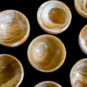 K&ouml;nnte beinhalten: Eine Ansammlung kleiner, runder Schalen aus durchscheinendem, goldbraunem Stein mit wei&szlig;en Adern. Die Schalen sind vor einem schwarzen Hintergrund angeordnet, der ihre polierten Oberfl&auml;chen und nat&uuml;rlichen Muster hervorhebt. Ideal zum Servieren kleiner Portionen.