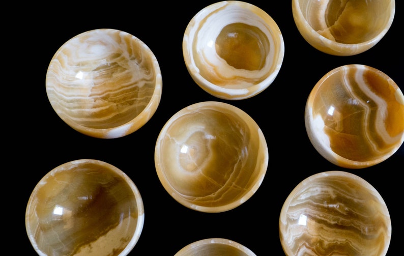 K&ouml;nnte beinhalten: Eine Ansammlung kleiner, runder Schalen aus durchscheinendem, goldbraunem Stein mit wei&szlig;en Adern. Die Schalen sind vor einem schwarzen Hintergrund angeordnet, der ihre polierten Oberfl&auml;chen und nat&uuml;rlichen Muster hervorhebt. Ideal zum Servieren kleiner Portionen.