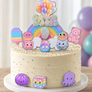 May include: A round cake with white frosting and colorful sprinkles, topped with a glittery cake topper featuring the name MIA, the number 9, a rainbow, balloons, and various cute cartoon characters including a bear, octopus, and cube.