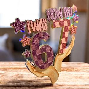 Puede incluir: Un adorno decorativo para tarta de cumpleaños con el texto "I WAS 6, NOW I'M 7". Los números y el texto tienen un patrón brillante a cuadros, con detalles de estrellas. El diseño está sostenido por manos de madera.