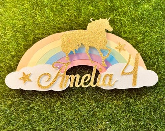 Op de afbeelding: Een witte en gouden glittertaarttopper met een regenboog, een eenhoorn, de naam Amelia en het nummer 4.