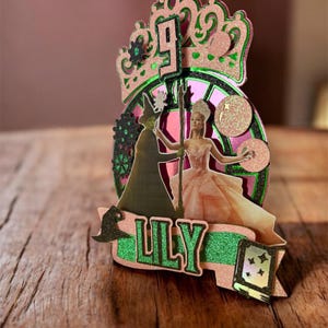 Könnte beinhalten: Ein mehrschichtiger Tortenaufsatz in Rosa und Grün. Das Design zeigt eine Krone, die Zahl 9 und zwei Figuren. Der Name "LILY" ist auf einem Banner zu sehen. Der Aufsatz steht auf einer Holzoberfläche.