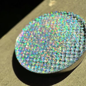 May include: A round, silver-coloured compact with a holographic, iridescent finish. The surface is covered in tiny, shimmering dots that create a rainbow effect.