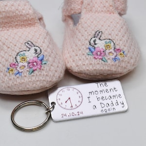 Könnte beinhalten: Ein silberner Schlüsselanhänger mit einem Zifferblatt und dem Text "The moment I became a Daddy again" und dem Datum "24.10.24". Der Schlüsselanhänger liegt neben einem Paar rosa Babyschuhe mit einem gestickten weißen Hasen.