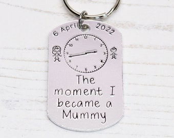 Pode incluir: Chaveiro de prata com uma etiqueta branca que diz "The moment I became a Mummy" e tem um mostrador de relógio com os ponteiros ajustados para 6 de abril de 2022.