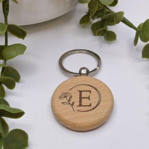 May include: Wooden keychain with a silver ring and a circular wooden charm. The charm has a floral design and the letter "E" engraved on it.