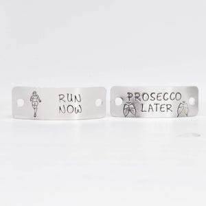 Op de afbeelding: Twee zilveren metalen labels met gegraveerde tekst. Het eerste label toont een hardlopend figuur en de tekst "RUN NOW". Het tweede label toont twee champagne glazen en de tekst "PROSECCO LATER".