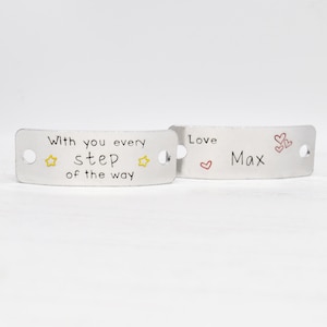 May include: Two silver metal tags with engraved text. The first tag says "With you every step of the way" with two yellow stars. The second tag says "Love Max" with a red heart and three smaller red hearts.