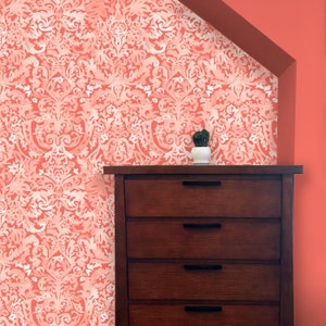 May include: A coral-colored wallpaper with a white floral pattern adorns a wall. A dark wooden dresser with four drawers and black handles sits below the wallpaper. A small cactus in a white pot is on top of the dresser.
