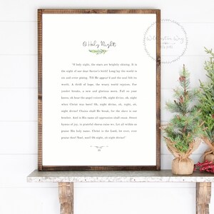 May include: A rectangular framed sign with a dark wood border featuring the text O Holy Night and the lyrics to the Christmas carol printed in black on a white background, accompanied by two small artificial pine trees in burlap bases.