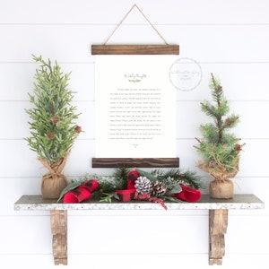 May include: A vertical wall hanging featuring a white paper print with black text titled Holy Night, secured between two dark brown wooden slats with a twine hanger. The print is flanked by two small artificial pine trees in burlap bases.