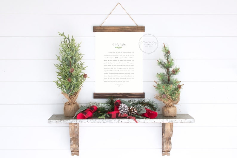 May include: A vertical wall hanging featuring a white paper print with black text titled Holy Night, secured between two dark brown wooden slats with a twine hanger. The print is flanked by two small artificial pine trees in burlap bases.