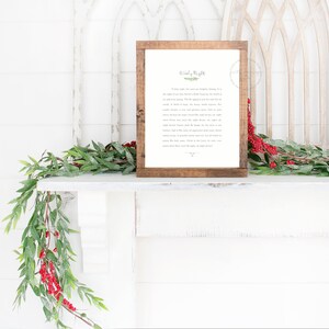 May include: A rectangular wooden frame containing a white print with the text Silent Night in black lettering, accompanied by a small green leaf illustration. The frame is positioned on a white mantelpiece adorned with green foliage and red berries.