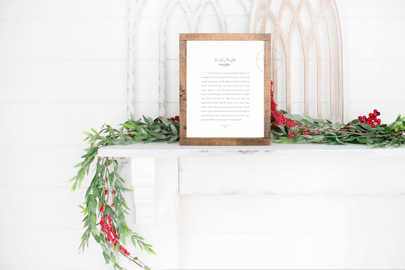 May include: A rectangular wooden frame containing a white print with the text Silent Night in black lettering, accompanied by a small green leaf illustration. The frame is positioned on a white mantelpiece adorned with green foliage and red berries.