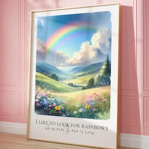 Pode incluir: Uma impressão artística emoldurada com um arco-íris vibrante sobre um vale verde com colinas e flores silvestres. A obra inclui o texto: "I like to look for rainbows whenever there is rain."