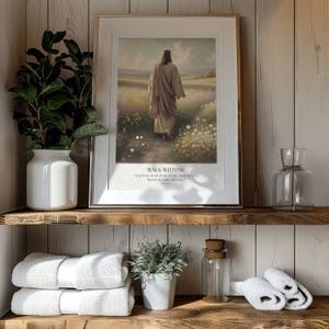 Puede incluir: Obra de arte enmarcada con el texto "WALK WITH ME" y una imagen de una persona caminando por un campo. La obra de arte se exhibe en un estante de madera con un jarr&oacute;n blanco, vegetaci&oacute;n, toallas blancas y una peque&ntilde;a planta en maceta. Un tarro de cristal transparente y una botella de cristal decorativa tambi&eacute;n est&aacute;n en el estante.
