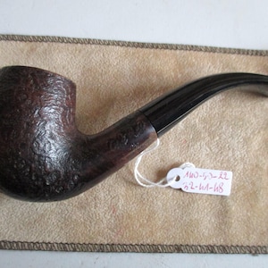 Pode incluir: Um cachimbo de tabaco castanho com uma tigela texturizada e uma haste preta curva. O cachimbo repousa sobre uma superfície bege. Uma pequena etiqueta com texto está presa ao cachimbo.