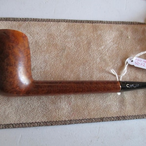Pode incluir: Um cachimbo de tabaco castanho com uma tigela curva e um longo caule. O cachimbo repousa sobre uma bolsa bege. O caule tem um bocal preto com a letra "C". Uma etiqueta com números está presa à bolsa.