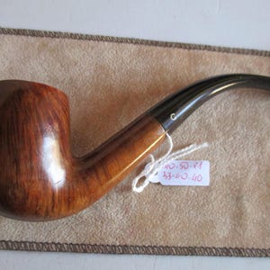 Pode incluir: Um cachimbo de tabaco de madeira com um bocal curvo preto. A tigela é castanha escura rica com grão de madeira visível. Uma pequena etiqueta está presa ao cachimbo. O cachimbo repousa sobre um pano bege.
