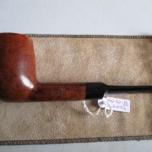 Pode incluir: Um cachimbo de tabaco castanho curvo com um bocal preto, sobre um saco bege. O cachimbo tem um acabamento brilhante e uma tigela arredondada. Uma etiqueta branca com texto está presa ao cachimbo.