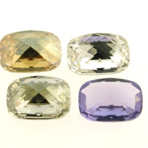 Pode incluir: Quatro pedras de vidro facetadas em diferentes cores: dourado, transparente, verde claro e roxo. As pedras são todas retangulares e têm uma forma ligeiramente arredondada.