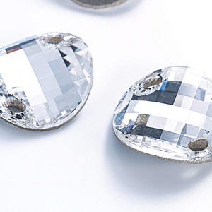 Op de afbeelding: Twee heldere kristallen naaisteentjes met een afgeronde, licht ovale vorm. Elk steentje heeft twee gaatjes om te naaien.