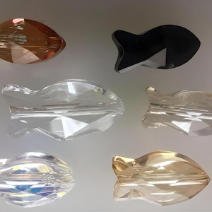 Op de afbeelding: Zes kristallen visbedels in verschillende kleuren, waaronder helder, iriserend, amber en zwart. De bedels zijn gefacetteerd en hebben een gat in het midden om op te hangen.