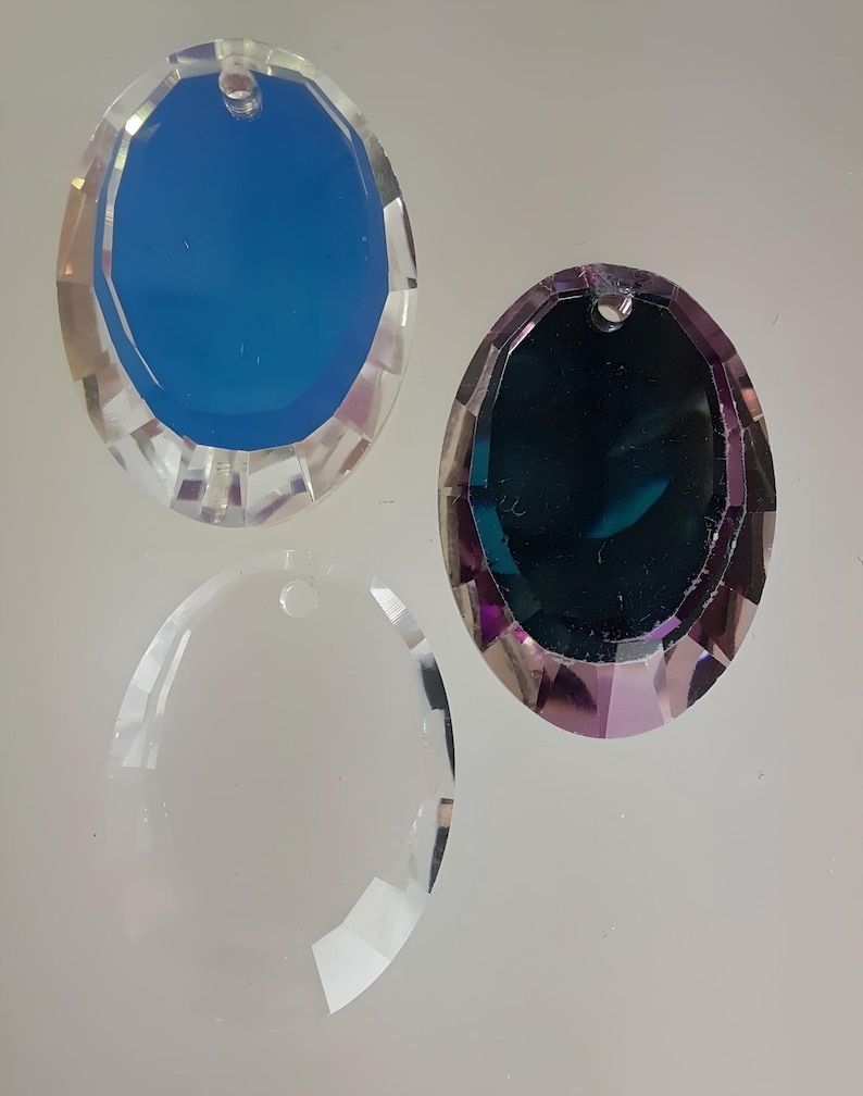 Puede incluir: Tres prismas de cristal de forma ovalada con un agujero en la parte superior para colgar. Los prismas son transparentes, azules y morados.