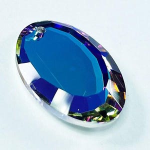 Puede incluir: Un gran prisma de cristal de forma ovalada con un acabado azul iridiscente. El prisma tiene un agujero en la parte superior para colgarlo.