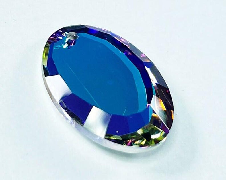 Puede incluir: Un gran prisma de cristal de forma ovalada con un acabado azul iridiscente. El prisma tiene un agujero en la parte superior para colgarlo.