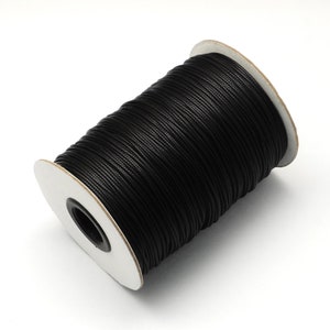 May include: A spool of black waxed cord, tightly wound and ready for use. The cord is a deep, solid black, and the spool has white cardboard sides. This versatile cord is suitable for various crafting and jewelry-making projects.