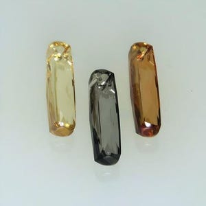 May include: Three faceted gemstones, one yellow, one brown, and one grey, are displayed on a white surface.