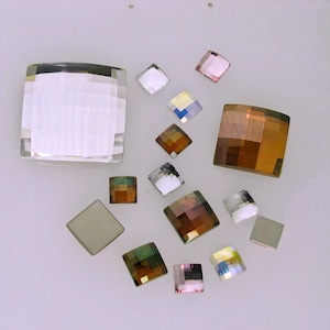 Può includere: Assortimento di strass quadrati in vetro di vari colori, tra cui trasparente, rosa, marrone e iridescente. Gli strass sono disposti su una superficie bianca.