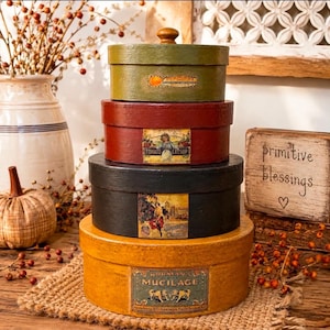 May include: A stack of four round wooden boxes with lids, featuring vintage-style labels. The top box is olive green, the second is deep red, the third is black, and the bottom box is mustard yellow, each displaying unique printed graphics.