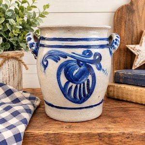 May include: A cylindrical ceramic crock with a cream-colored glaze and two side handles. The exterior features hand-painted cobalt blue floral motifs and horizontal bands. The vessel has a wide opening and a sturdy base, showcasing a rustic finish.