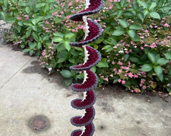 Könnte beinhalten: Eine gehäkelte Hängedeko mit Spiraldesign. Die Deko ist aus burgunderfarbenem und grauem Garn gefertigt und hängt an einer weißen Schnur.
