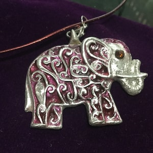 May include: A silver elephant pendant with a pink and silver swirl design and a small amber-colored stone for an eye. The pendant is hanging from a brown cord.