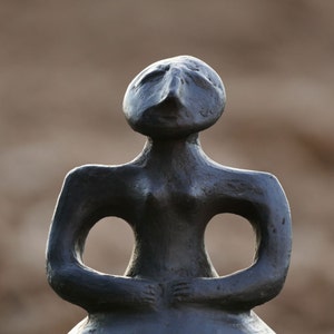 Pode incluir: Uma escultura em bronze de uma figura sentada com uma cabe&ccedil;a arredondada e tra&ccedil;os faciais alongados. Os bra&ccedil;os da figura est&atilde;o dobrados, com grandes aberturas onde os bra&ccedil;os encontram o torso. A escultura tem um acabamento liso e polido.