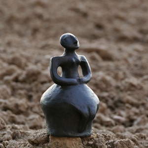 Pode incluir: Uma escultura cinza escuro de uma figura com os bra&ccedil;os cruzados e uma saia arredondada. A est&aacute;tua est&aacute; colocada sobre um pequeno peda&ccedil;o de madeira e assenta sobre um leito de terra. A figura olha para cima.