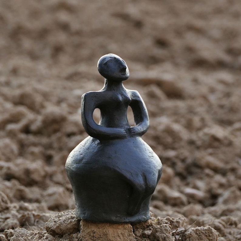 Pode incluir: Uma escultura cinza escuro de uma figura com os bra&ccedil;os cruzados e uma saia arredondada. A est&aacute;tua est&aacute; colocada sobre um pequeno peda&ccedil;o de madeira e assenta sobre um leito de terra. A figura olha para cima.