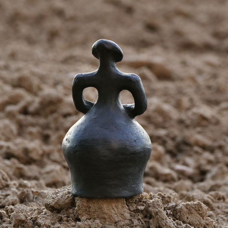 Pode incluir: Uma escultura de cer&acirc;mica cinza escuro de uma figura humana estilizada. A figura tem um corpo arredondado, bra&ccedil;os com grandes aberturas e uma cabe&ccedil;a pequena. A escultura est&aacute; sobre um fundo desfocado e terroso.