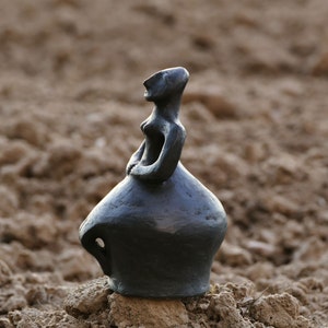 Pode incluir: Uma escultura cinza escuro de uma figura sentada com uma saia arredondada e os bra&ccedil;os cruzados sobre o peito. A figura tem um pesco&ccedil;o fino e uma cabe&ccedil;a inclinada. A escultura &eacute; colocada em uma pequena pilha de terra.