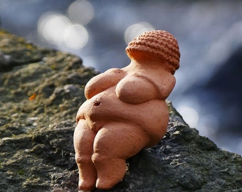 Peut inclure: Une figurine en terre cuite d'une femme au corps rond et à la tête texturée. La figurine est assise sur un rocher.
