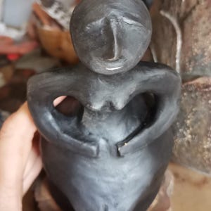 Pode incluir: Uma escultura cinza escuro de uma figura sentada com os bra&ccedil;os cruzados sobre o peito. A figura tem uma cabe&ccedil;a arredondada e um acabamento liso e fosco. A escultura &eacute; segurada em uma m&atilde;o.