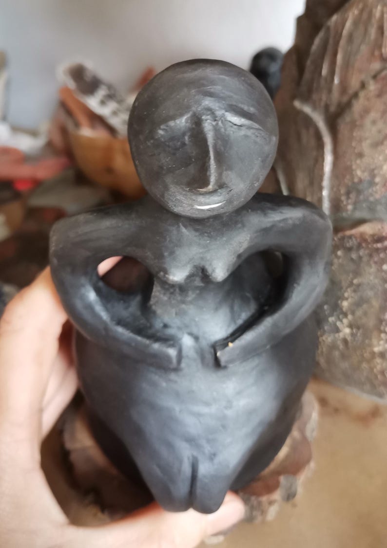 Pode incluir: Uma escultura cinza escuro de uma figura sentada com os bra&ccedil;os cruzados sobre o peito. A figura tem uma cabe&ccedil;a arredondada e um acabamento liso e fosco. A escultura &eacute; segurada em uma m&atilde;o.