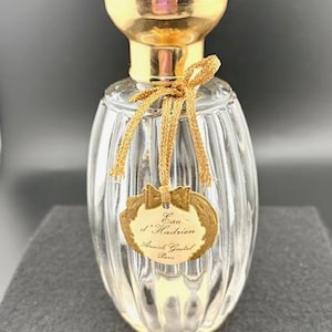 May include: A clear glass perfume bottle with a gold-colored cap and a gold-colored label. The bottle has a ribbed design and is tied with a gold ribbon. The label reads "Eau d'Hadrien Annick Goutal Paris."