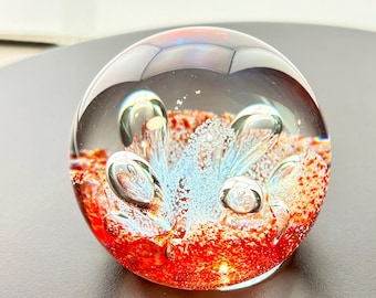 May include: A spherical glass paperweight featuring a clear outer layer with an internal design of vibrant red speckled glass and suspended air bubbles. The center contains a cluster of light blue glass accents and small white particles.